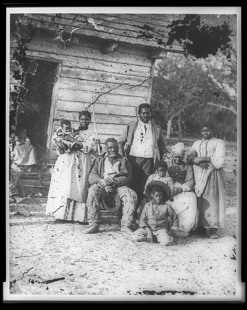 Black-and-white photograph of eight Black people sitting or standing as they pose outside a small, simple building