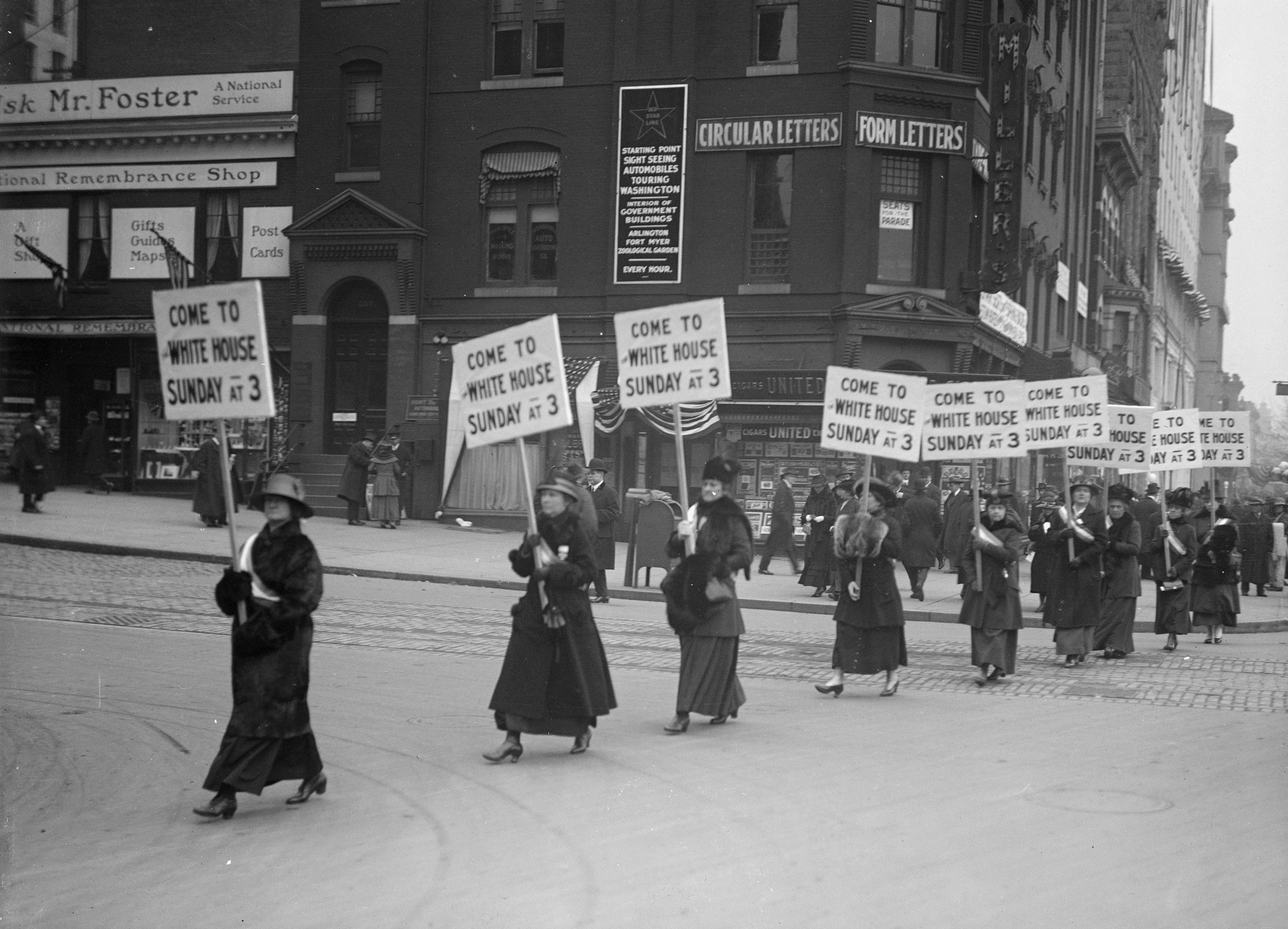 Black-and-white photograph of a line of women crossing a street while holding signs that read, “Come to White House Sunday at 3”