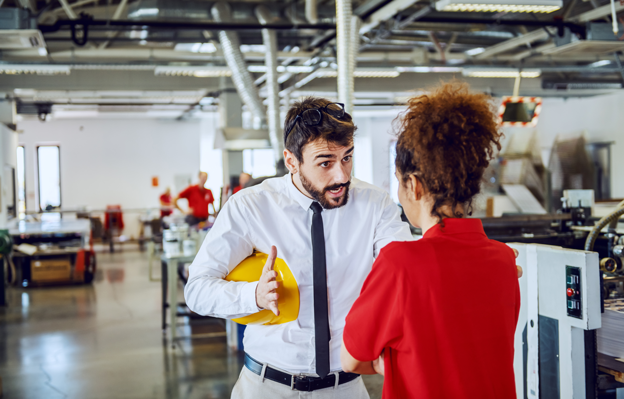 Man talking to woman in a warehouse environment. His body language and hers suggest he is giving harsh criticism.