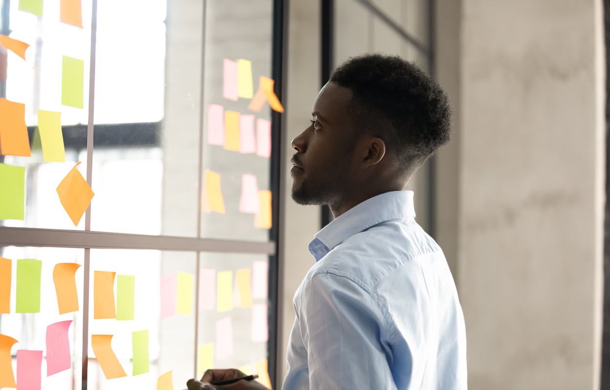 Man looking at a window, each pane with a cluster of post-it notes. He is using the window and notes as an organizational strategy.