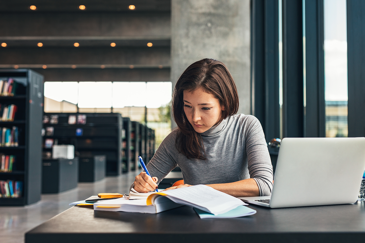 A young woman sits at a table in a library with a book, paper, and pencil.