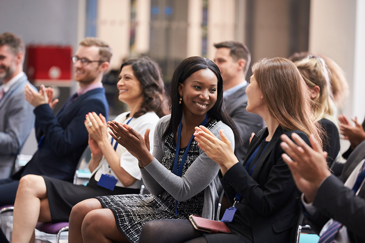 An image of an audience clapping and smiling as they listen to a speaker.