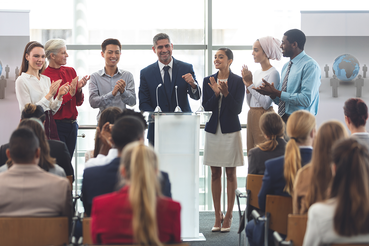 A group of professionally-dressed people stand at a podium in front of an audience.