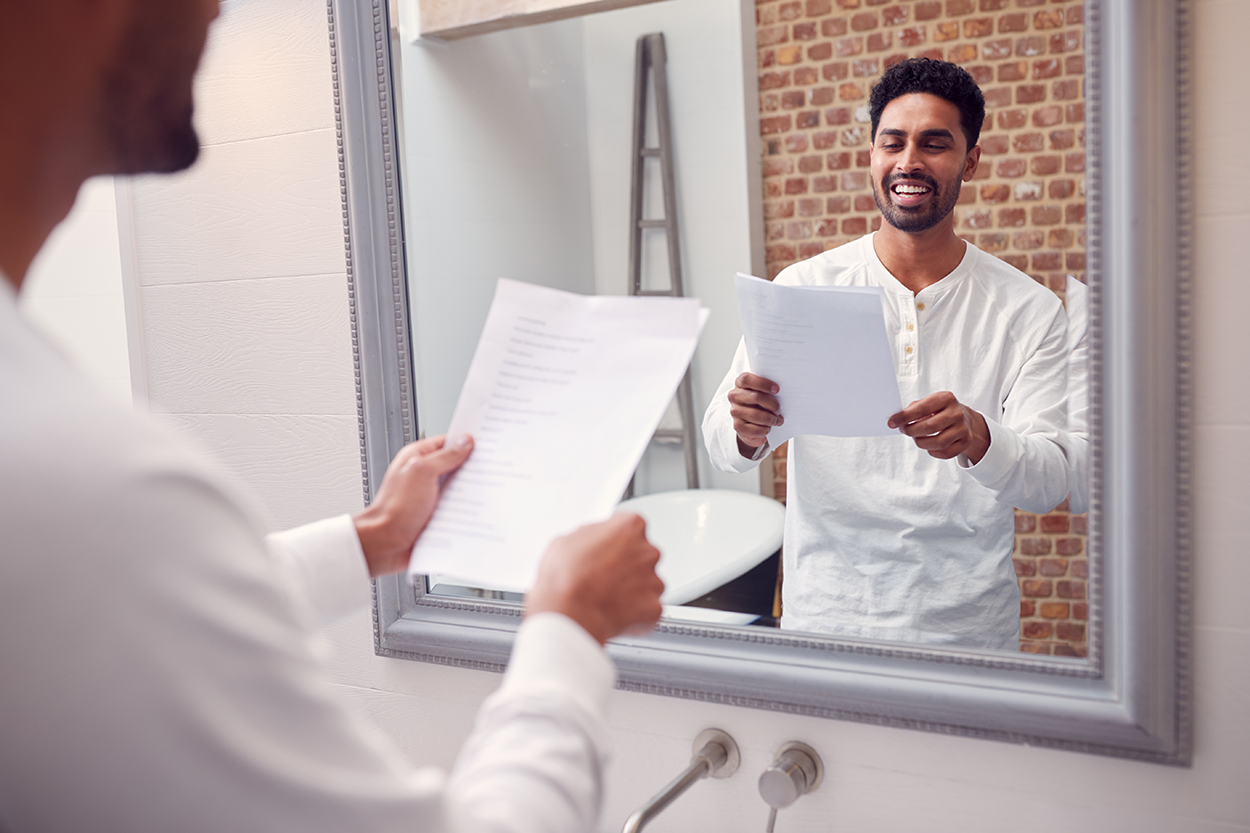 An image of a man holding a piece of paper and rehearsing his speech in a mirror.