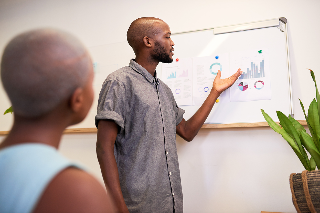 A man stands in front of a board with several diagrams on it, gesturing while he speaks to an audience.