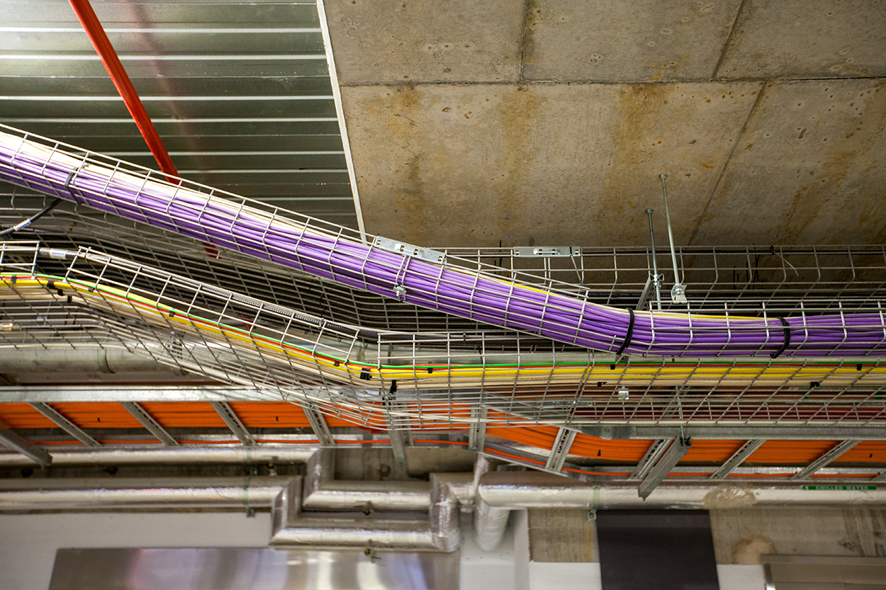 A complex network of cables and wires suspended from the building’s ceiling, with cables grouped into colorful bundles and fastened to metal racks or trays. These racks, which can be horizontal or sloping, are attached to the ceiling using metal brackets and wires, forming a grid-like framework.