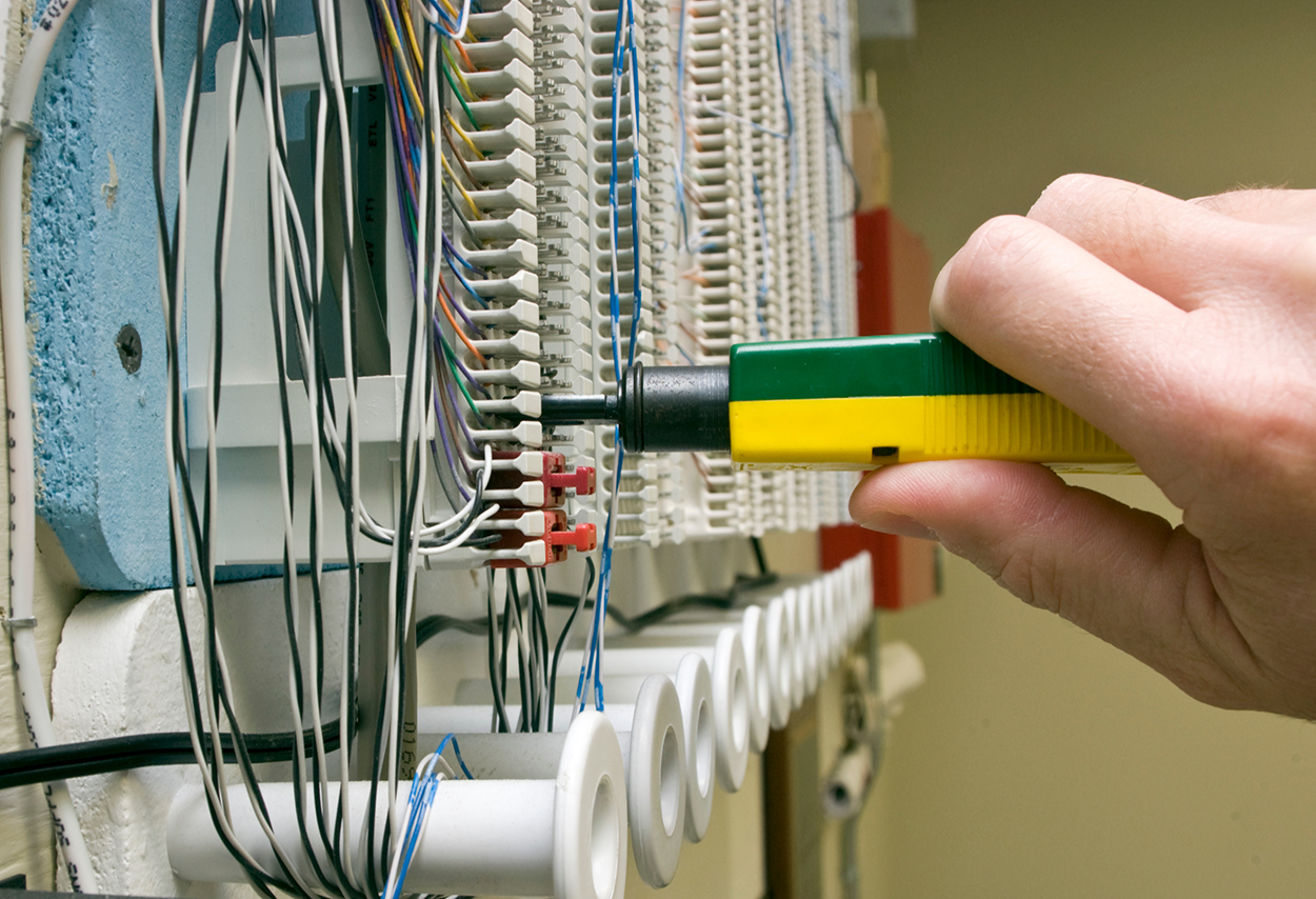A person is using a punch-down tool to secure wires into a punch-down block. The block contains multiple-colored wires that are already connected and arranged in order. In the background, a wall-mounted panel filled with numerous cables indicates a structured cabling system.
