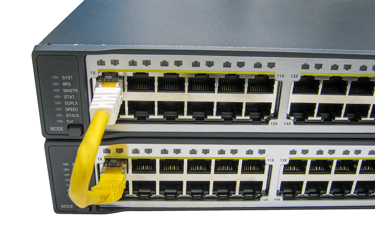 Two rectangular network switches are stacked one above the other and have several Ethernet ports arranged in two horizontal rows. These ports, which are rectangular sockets, are numbered from 1 to 17 in each switch. An Ethernet loopback plug is connected between the switches. This plug consists of a thick cable with two connectors of different colors—one connector is inserted into Port 1 of the top switch, and the other connector into Port 1 of the bottom switch. On the left side of each switch are indicator lights labeled ‘SYST’, ‘RPS’, ‘MASTR’, ‘STAT’, ‘DUPLX’, ‘SPEED’, ‘STACK’, and ‘P o E’ from top to bottom. Beneath the indicators is a round button labeled ‘MODE’. ‘1x’ is mentioned to the left of Port 1 and ‘11x’ to the right of Port 12.
