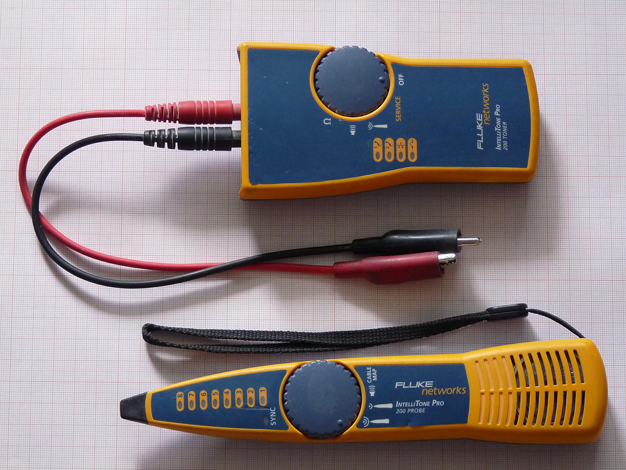 A Fluke Networks IntelliTone Pro 200 toner-and-probe kit consists of two devices: 1) Toner: This is a compact, wide unit with a rotary dial on one side and two options below the dial: ‘Service’ and ‘Tone’. Next to the dial, there are three icons: speaker, signal, and ohm. Two output cables—a red one and a black one—extend from the bottom of the toner, each ending in an alligator clip. 2) Probe: This is a slim, pen shaped device with a central rotary dial and eight numbered signal level buttons (1 through 8) arranged vertically above the dial. SYNC is mentioned above the dial. Below the dial, there are three icons: a speaker and two signals. The probe tapers from a wider base down to a pointed tip and includes a wrist strap for easy handling. When the toner is active, the probe picks up and audibly indicates the tone signal along a cable.
