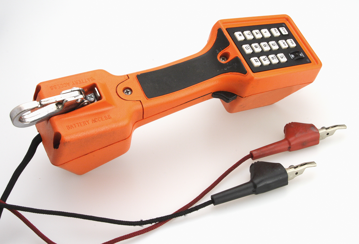 A butt set is a tool that resembles a telephone handset with a handle in the middle. The top side features a keypad with number buttons from 0 to 9, as well as symbols like asterisk, hash, and more. On the other side of the device, there's a connector or alligator clip, and below it, a point where two wires emerge—one red and one black, each with metal endings.