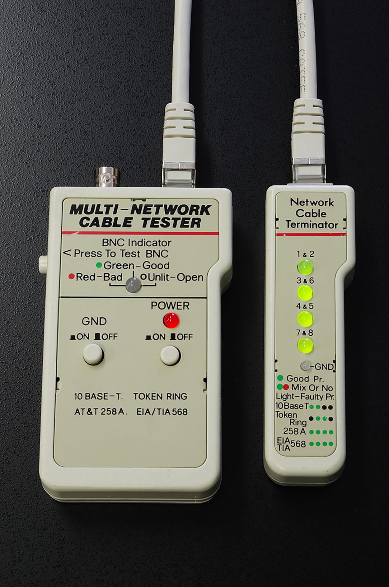 A network cable tester consists of two rectangular handheld units: 1) The main unit (on the left) is labeled ‘MULTI-NETWORK CABLE TESTER’. It has a BNC indicator section near the top with the instructions ‘Press To Test BNC’. Green Dot Green—Good represents the green light symbol for a good connection. Red Dot Red—Bad represents a red light symbol for a bad connection. Unshaded Dot Unlit—Open represents no light symbol for an open circuit. Beneath this section are two toggle switches labeled ‘GND ON/OFF’ and ‘POWER ON/OFF’, each with a corresponding round button. The power button is lit red, indicating that the unit is powered on. At the bottom of the unit, supported cabling standards are listed: ‘10 BASE-T’, ‘TOKEN RING’, ‘AT&T 258A’, and ‘EIA/TIA 568’. 2) The remote unit (on the right) is labeled ‘Network Cable Terminator’. Connected to the main unit via an Ethernet cable. It has four pairs of small round lights, each corresponding to a pair of Ethernet wires: 1 & 2, 3 & 6, 4 & 5, 7 & 8. Additionally, there is a single light labeled ‘GND’. Below the indicator lights, a legend explains the test result color codes: 1) Green dot: Good Pair, 2) Green and Mixed dot: Mix Or No Light—Faulty Pr, 3) 10 Base T: green dot, green dot, black dot, and black dot, 4) Token Ring: black dot, green dot, green dot, and black dot, 258 A: green dot, green dot, green dot, and green dot, 5) EIA 568: green dot, green dot, green dot, and green dot, 6) TIA