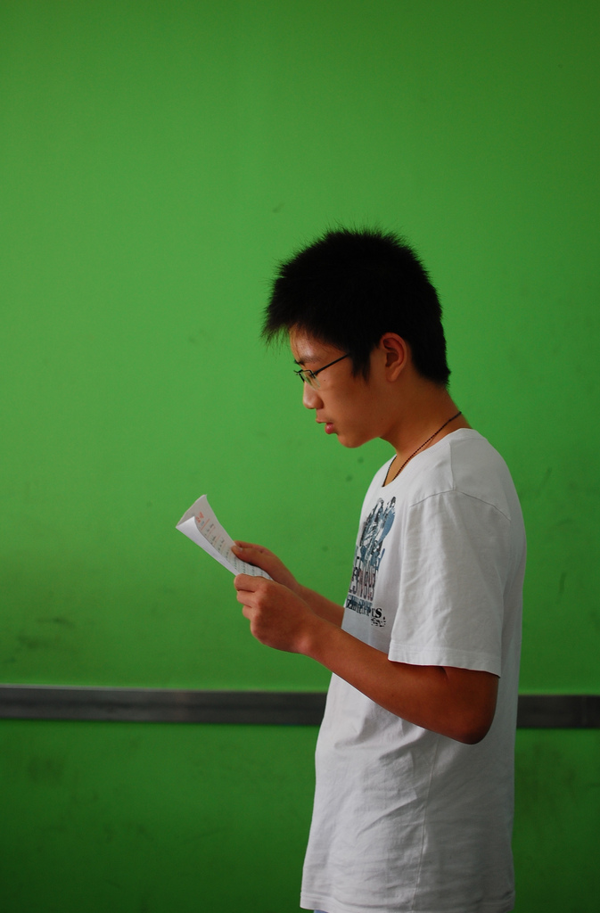 A young man in a white t-shirt and glasses reads from a piece of paper.