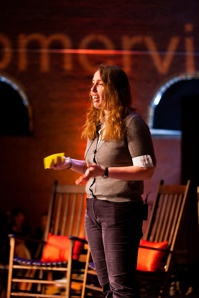 A woman standing on a stage holds cue cards in her hand and wears a wireless microphone. She is speaking to an audience and gesturing with her hands.
