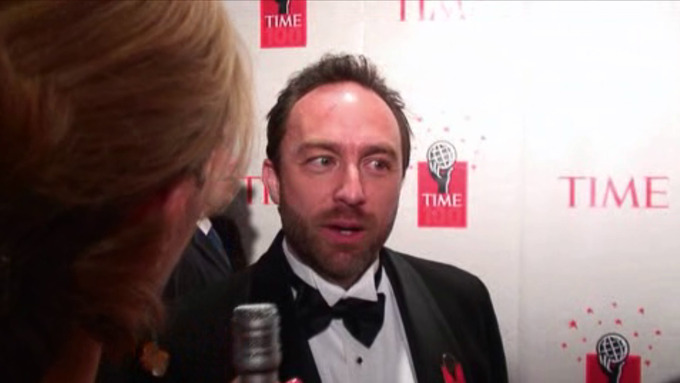 A photograph of a man in a tuxedo speaking to a woman holding a microphone. As he pauses his speech, he looks to one side.