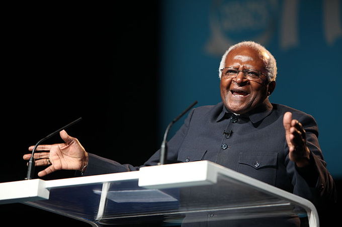 A man stands at a podium speaking into a microphone and holds his arms out wide in a gesture.