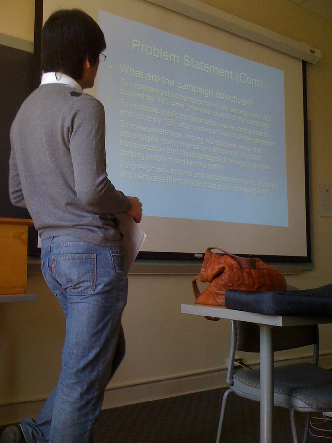 An image of a speaker using a projector screen to present multimedia content to the audience.