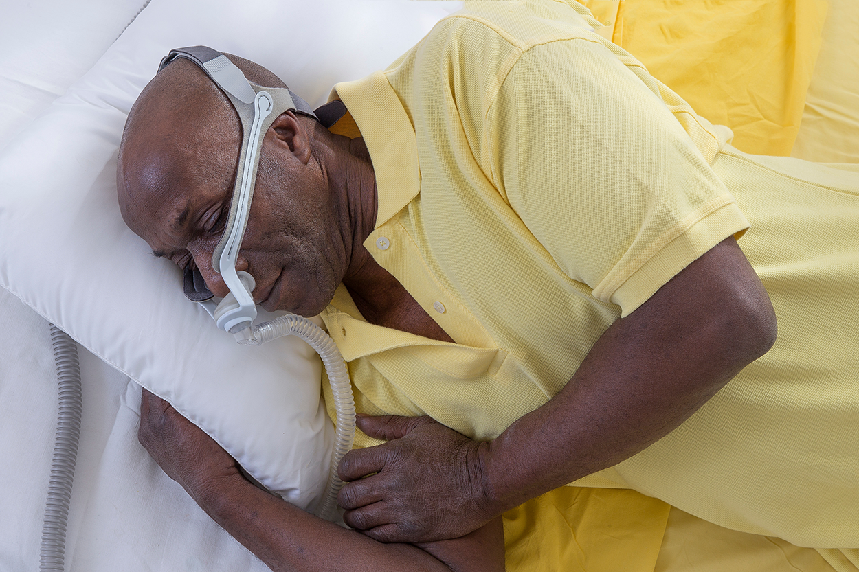 Man lying in bed on this right side with his head on a pillow.  On his head is a CPAP machine that is allowing him to sleep.