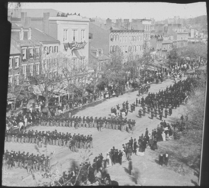 President Abraham Lincoln's funeral procession on Pennsylvania Avenue on April 19, 1865. A procession of several hundred people moves down a street lined with tall buildings.