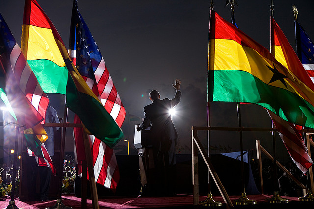 An image of President Barack Obama wearing a suit and surrounded by flags while standing at a podium. His arm is raised as he delivers a speech.