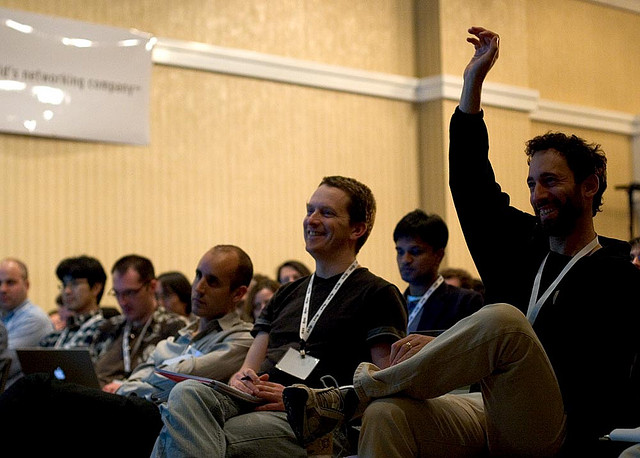 An image of an audience of people is shown. One of the audience members has his hand raised.