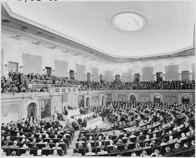 An old photograph of a man in a suit standing in the House Chamber of the United States Capitol Building delivering a speech to an audience.