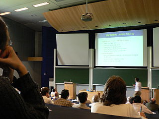 A large lecture hall is shown with a person delivering a lecture using a projected slide. There is an audience sitting in chairs listening to the lecture.