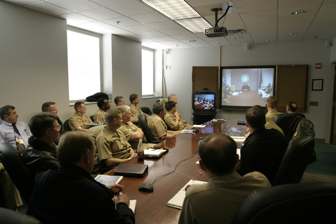 Military personnel seated around a large boardroom table participate in a meeting. There is a large screen on the far wall where a projected video feed of another board room is displayed.