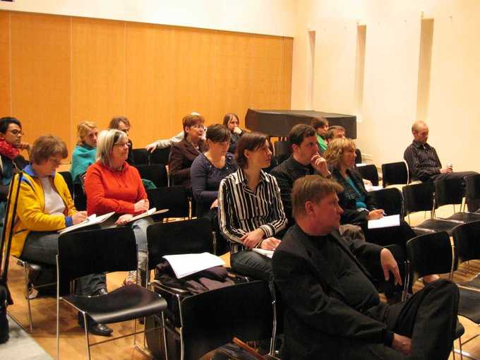 An audience is shown sitting in chairs listening to a speaker in a small room.