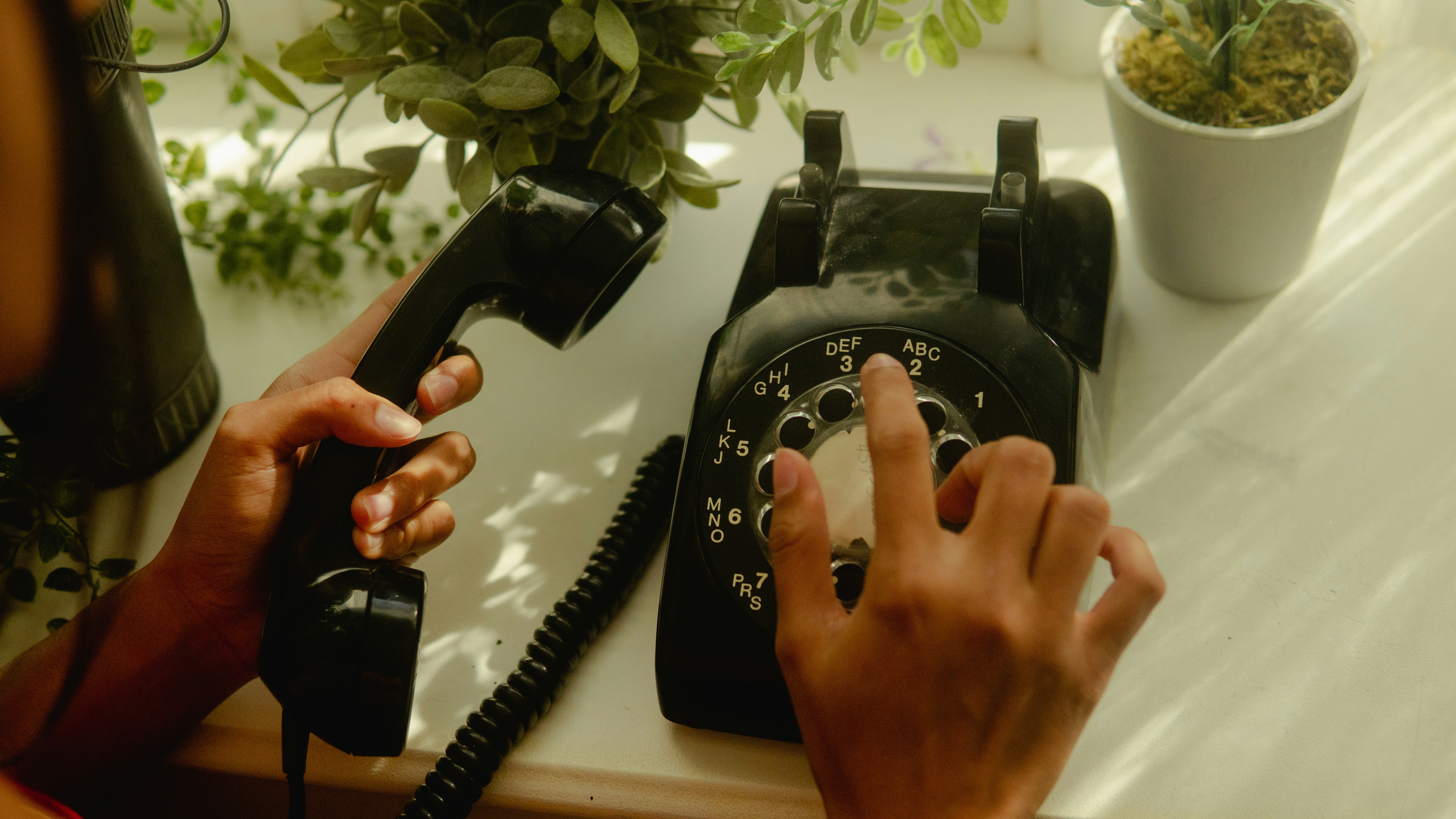 A person holding the receiver of a rotary dial phone with one hand while dialing a number with the other. The rotary phone has a circular dial with finger holes arranged in a ring, each corresponding to a number from 1 to 9 and 0. The handset is connected to the top of the rotary phone with a coiled cord.