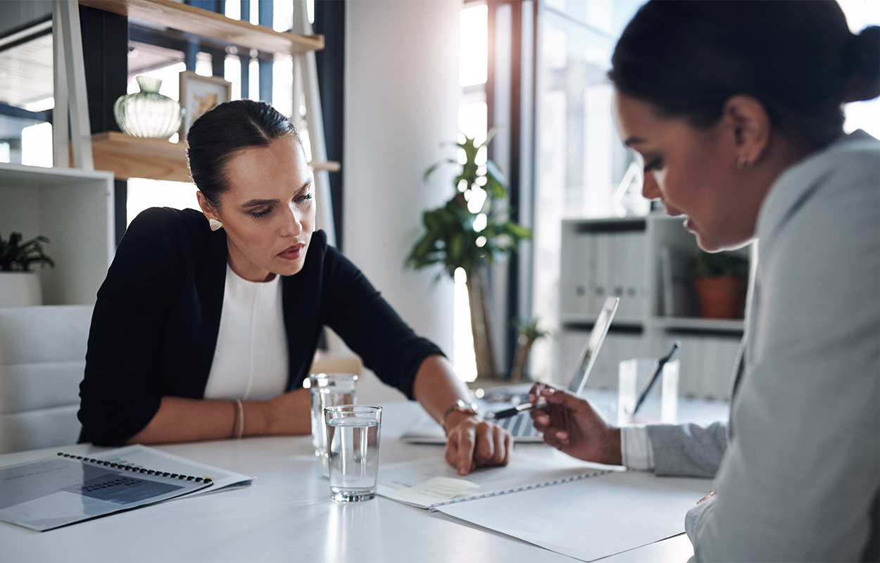 An image two employees in a professional office setting. One employee is discussing paperwork with the other.