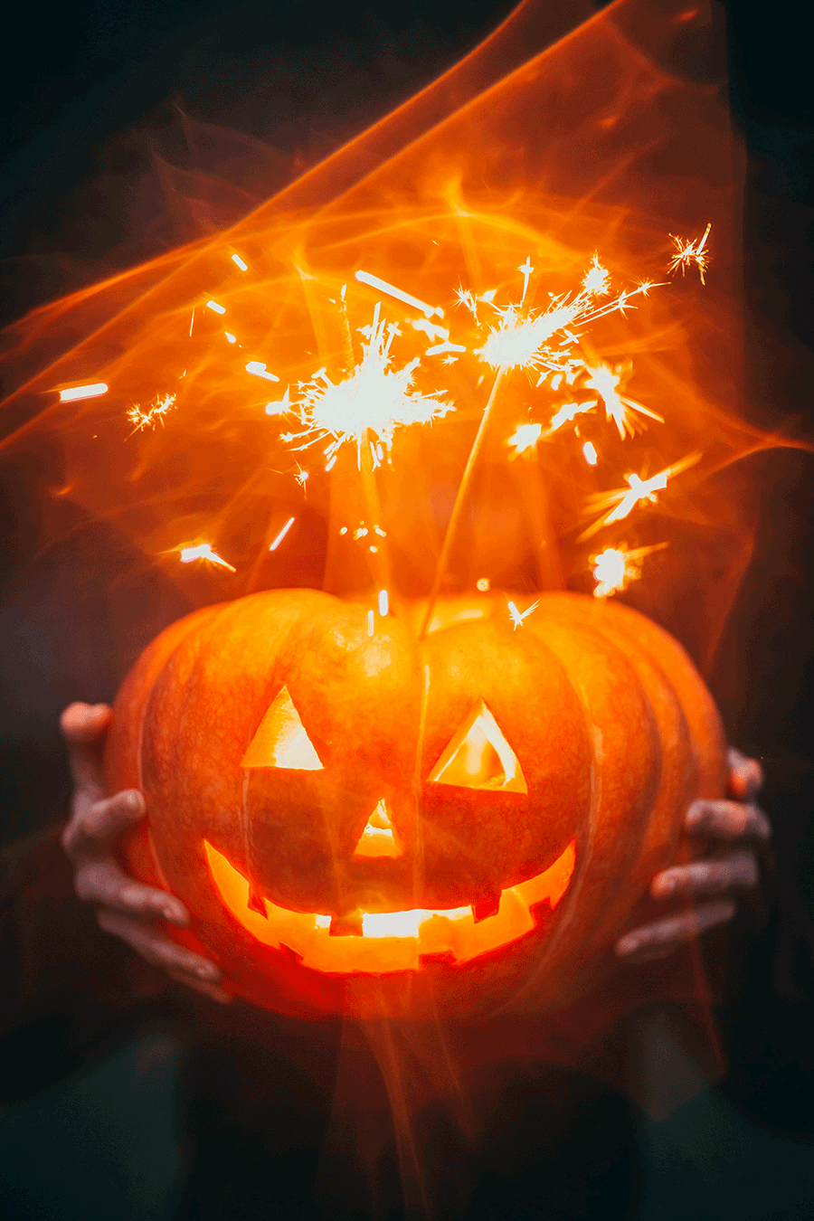 A person holds a carved Jack-O-Lantern with orange light emanating from its face and a pair of sparklers.