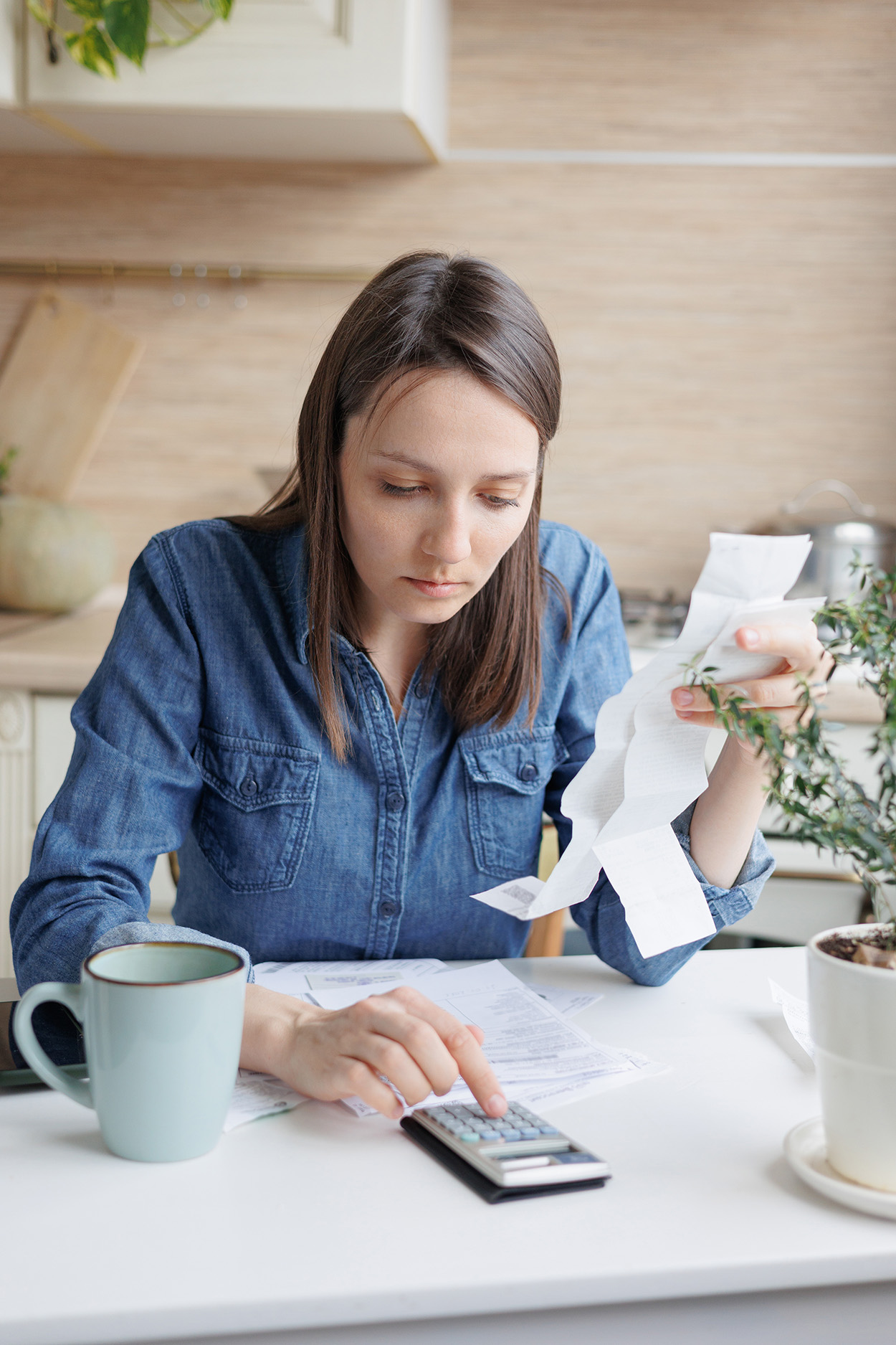 This image captures a thoughtful individual reviewing receipts and using a calculator, representing personal finance management. It portrays a common scenario of budgeting, tracking expenses, or planning finances. The setting, likely at home, conveys the importance of staying on top of financial responsibilities in a comfortable and focused environment. The coffee mug and casual attire emphasize a personal, everyday approach to handling financial tasks.