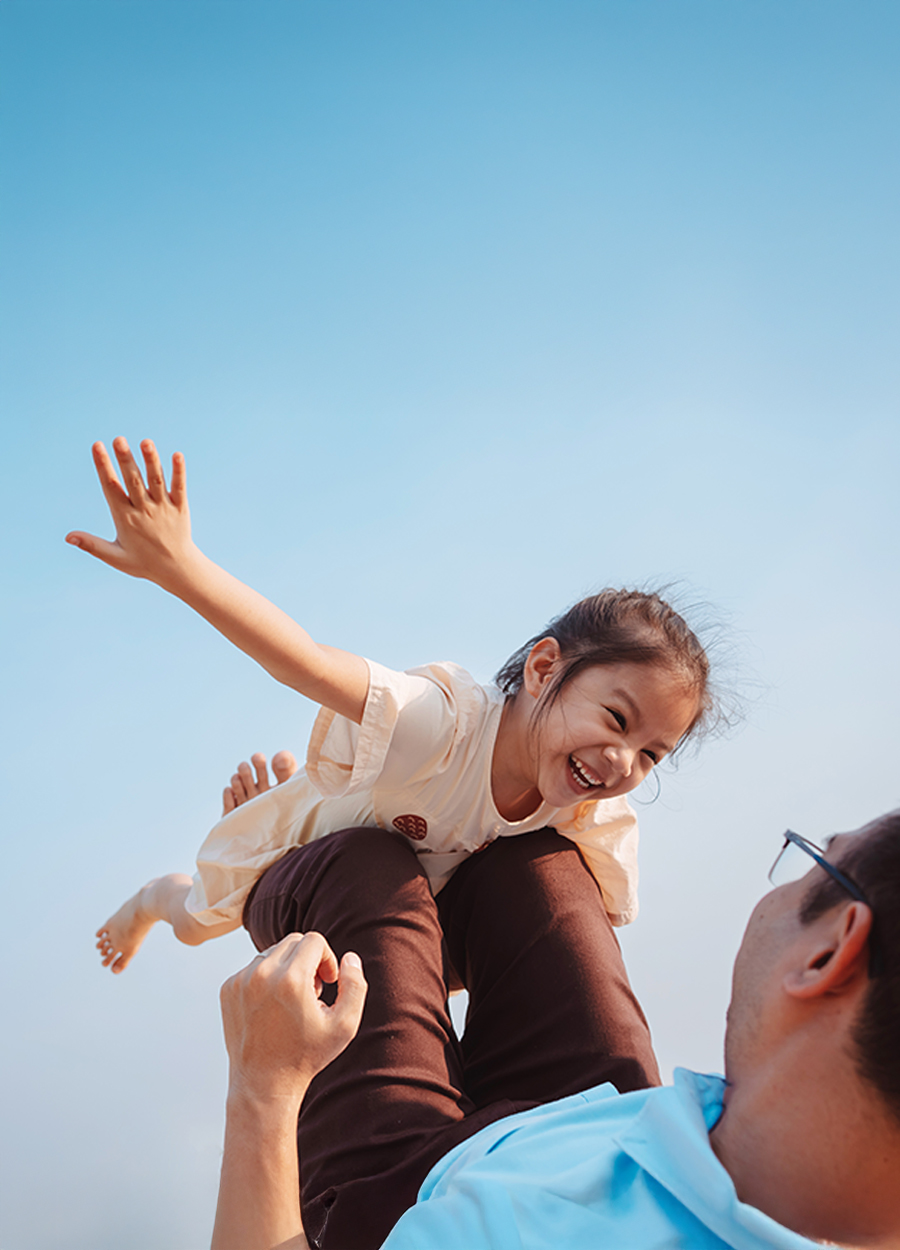 The image captures a joyful moment between a father and his young daughter outdoors. The father, wearing glasses and a short-sleeved shirt, is lying on his back with his legs bent, lifting his daughter into the air with his legs supporting her. The daughter, dressed in a light-colored dress, is extending her arms wide with a big smile on her face as if pretending to fly. The background consists of a clear sky, emphasizing the bright and cheerful atmosphere of the scene.