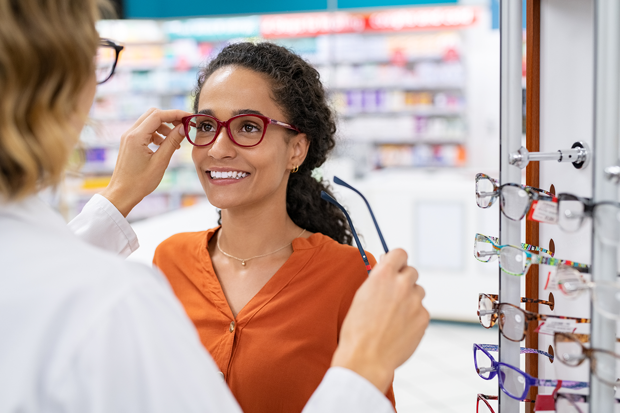 The image shows a woman trying on a pair of eyeglasses at an optical store. She is smiling as an optician dressed in a lab coat assists her in adjusting the frames. The woman holds another pair of glasses in her hand while standing in front of a display rack filled with various eyeglass frames. The background, slightly blurred, features shelves stocked with products, indicating a retail or pharmacy setting. The scene captures the moment of selecting new eyewear, likely as part of a vision care appointment or insurance-covered purchase.