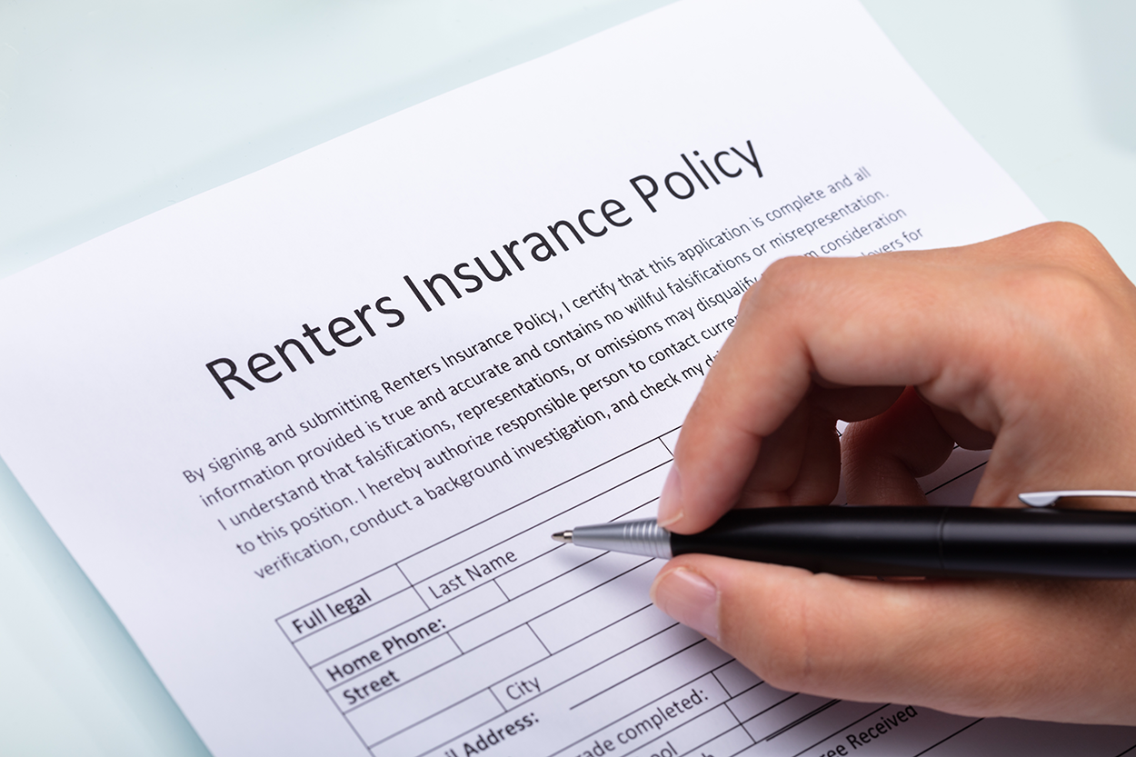 The image shows a close-up of a person filling out a renters insurance policy form. The form contains sections for personal information such as full legal name, home phone number, street address, and email address. A hand is holding a pen, poised to write on the document. The text on the form emphasizes the importance of providing accurate information and mentions verification and background checks. The setting suggests an administrative or personal documentation process related to obtaining renters insurance.