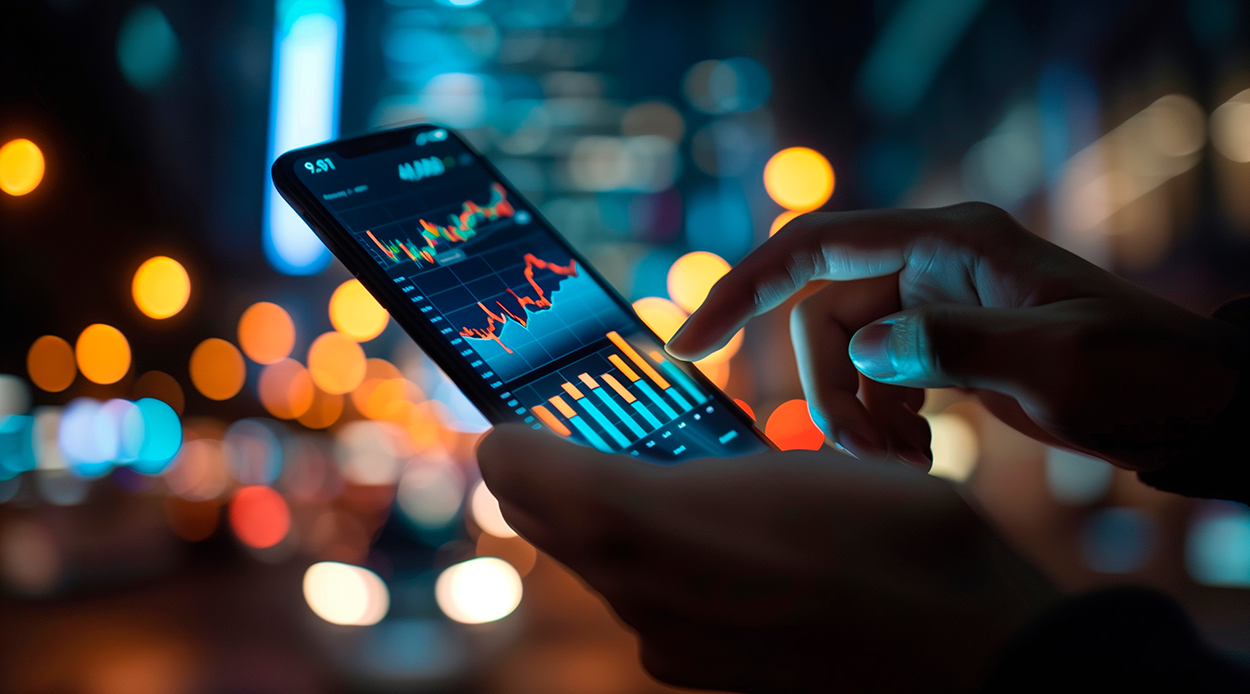 This image shows a person using a smartphone to monitor stock market data, likely engaging in real-time trading or investment tracking. The phone screen displays financial charts, including candlestick patterns and bar graphs, indicating market fluctuations and trends. The background is blurred with glowing city lights, suggesting an urban setting, possibly at night. The scene conveys a sense of financial engagement, technology-driven investing, and the fast-paced nature of stock trading in the digital age.
