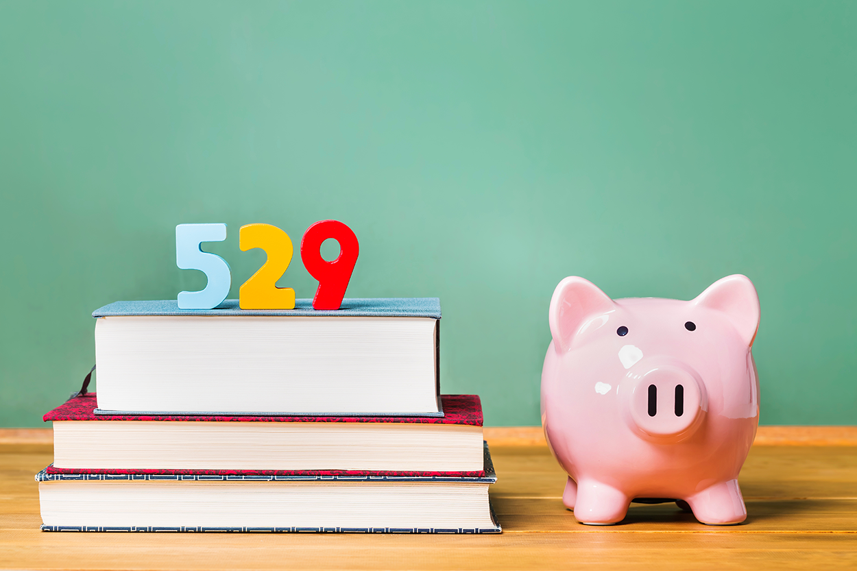 This image visually represents the concept of a 529 savings plan, which is a tax-advantaged account designed for educational expenses. It features a stack of books with the number “529” placed on top, symbolizing education and savings. Beside the books, a piggy bank represents financial planning and investment for future education costs. The background consists of a chalkboard, reinforcing the theme of learning and education.