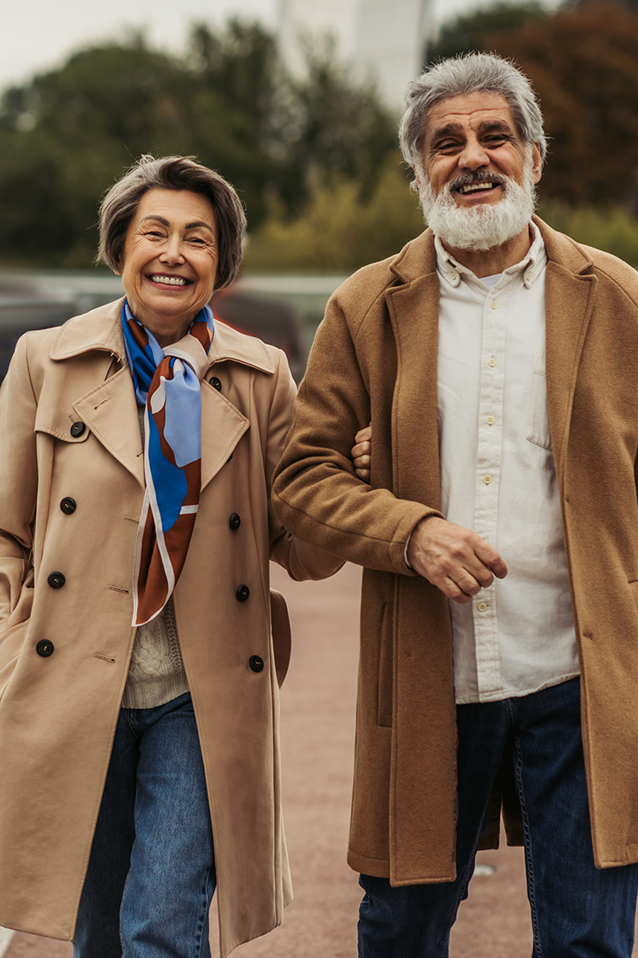A smiling older couple walks arm in arm, dressed in stylish coats, enjoying a leisurely stroll outdoors.