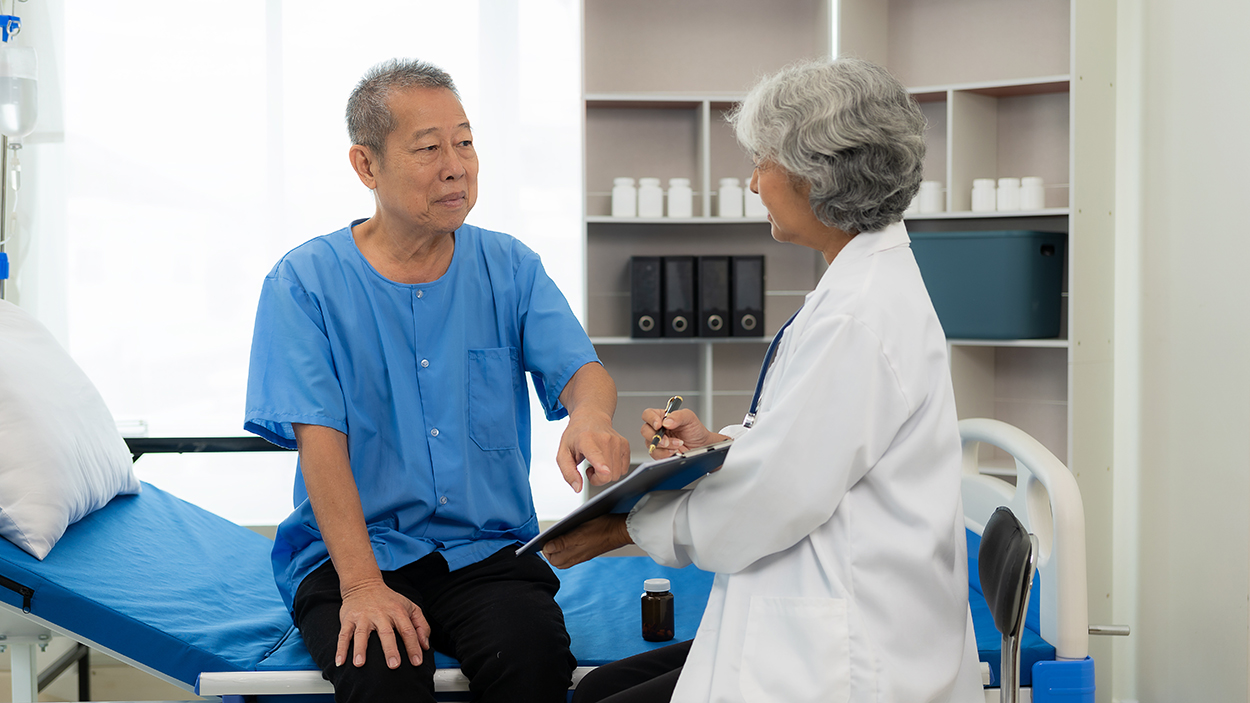 An elderly patient in a hospital gown sits on a bed, discussing his condition with a senior doctor holding a clipboard in a medical examination room.