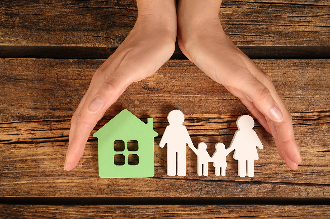 A pair of hands positioned protectively over wooden cutouts of a house and a family of four, symbolizing home and family security.