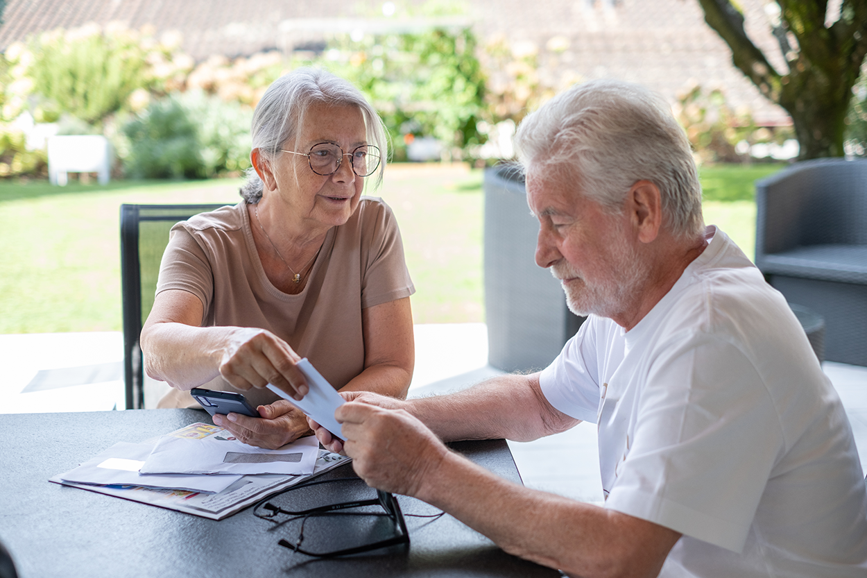 An elderly couple is sitting at an outdoor table, engaged in reviewing documents. The woman, wearing glasses and a short-sleeved shirt, is holding a smartphone and handing a piece of paper to the man. The man, who has a beard and mustache, is attentively receiving the document. Various papers, envelopes, and a pair of eyeglasses are scattered on the table. The background features outdoor furniture and trees, suggesting a relaxed setting, possibly in a backyard or patio.