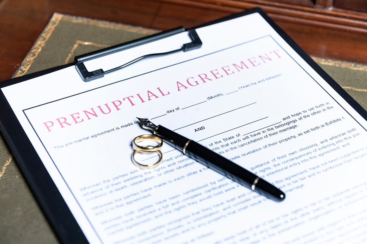 A prenuptial agreement document on a clipboard, with a fountain pen and two wedding rings placed on top. The document has blank spaces for names and dates, indicating it is ready to be filled out.