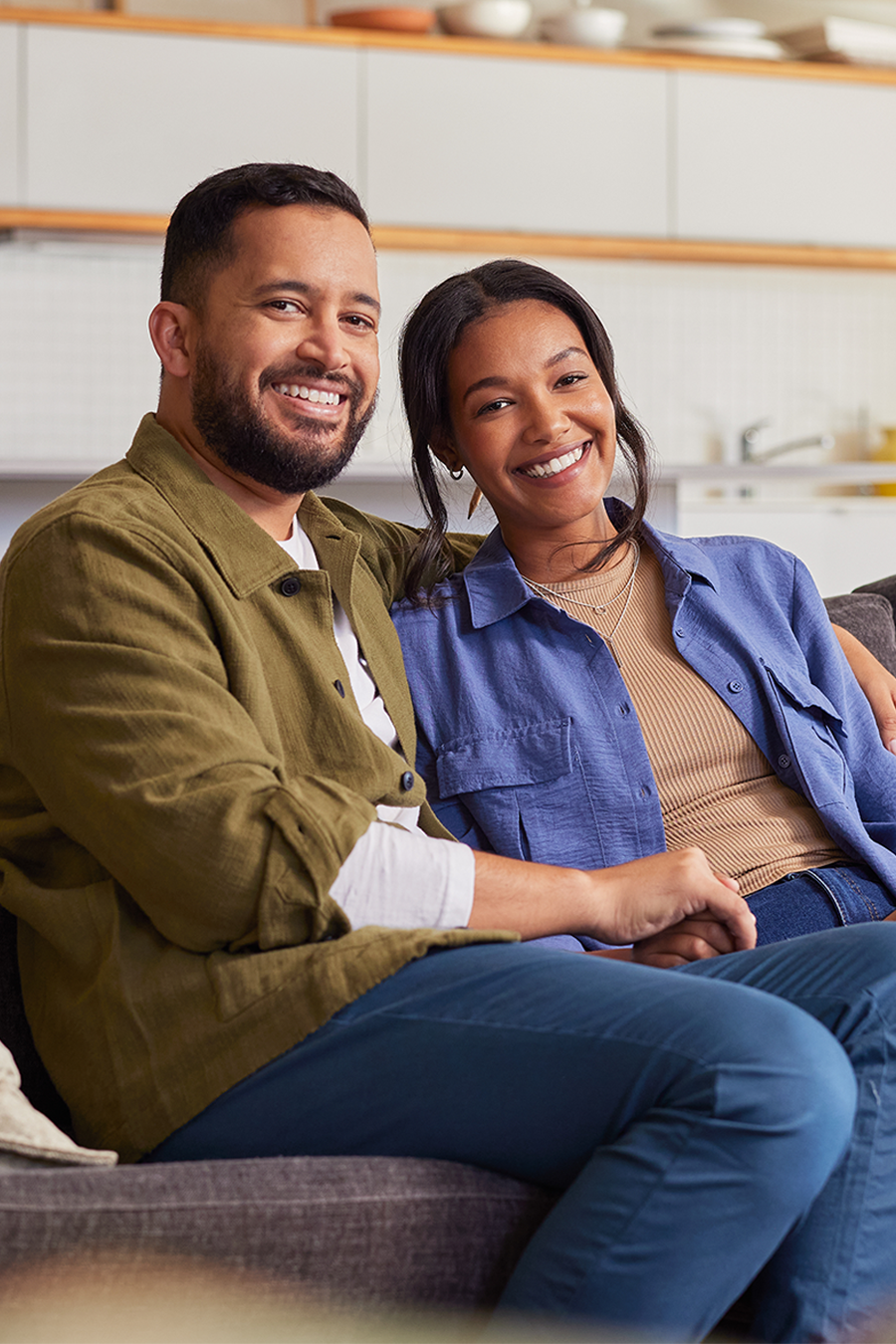 A smiling couple sits closely together on a comfortable couch in a well-lit, modern living room. The man has his arm around the woman, and they appear happy and relaxed. The background features a contemporary kitchen with shelves, kitchenware, and a large window that lets in natural light.