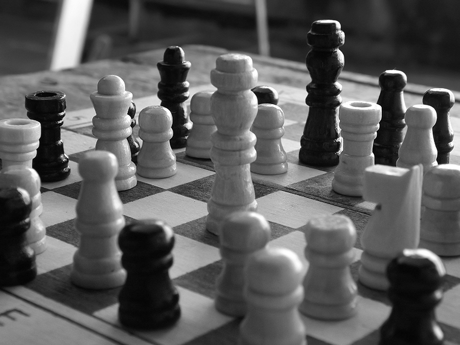 A close-up black-and-white photo of a chessboard with many wooden pieces in play.