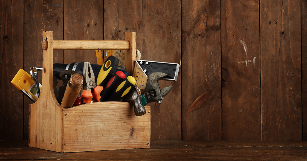 A wooden toolbox filled with assorted hand tools in front of a rustic wooden wall.
