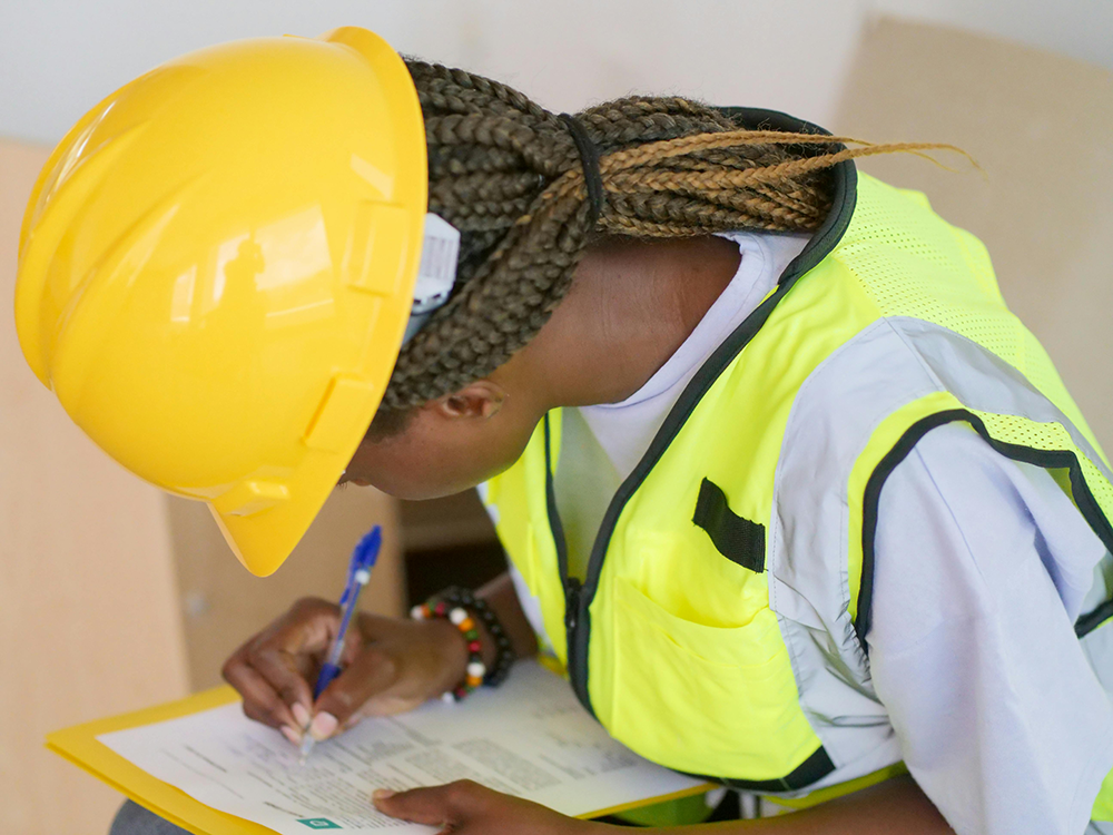 A person in a safety vest and hard hat filling out a form on a clipboard.