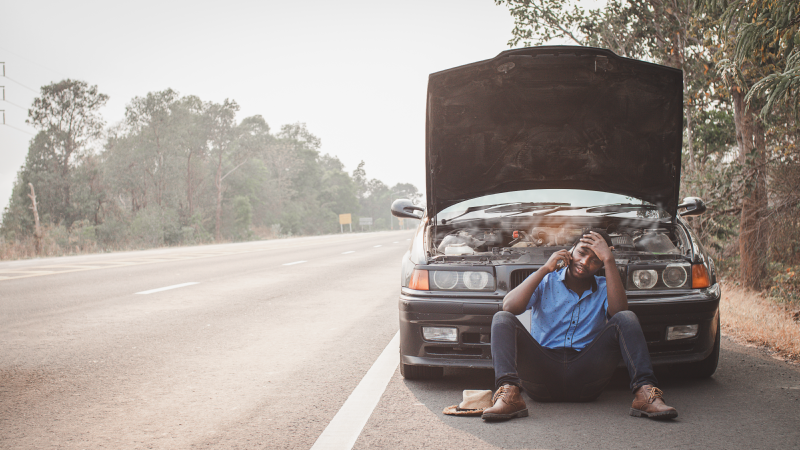 A man sitting on the roadside in front of a car with the hood up, talking on a phone and looking frustrated.