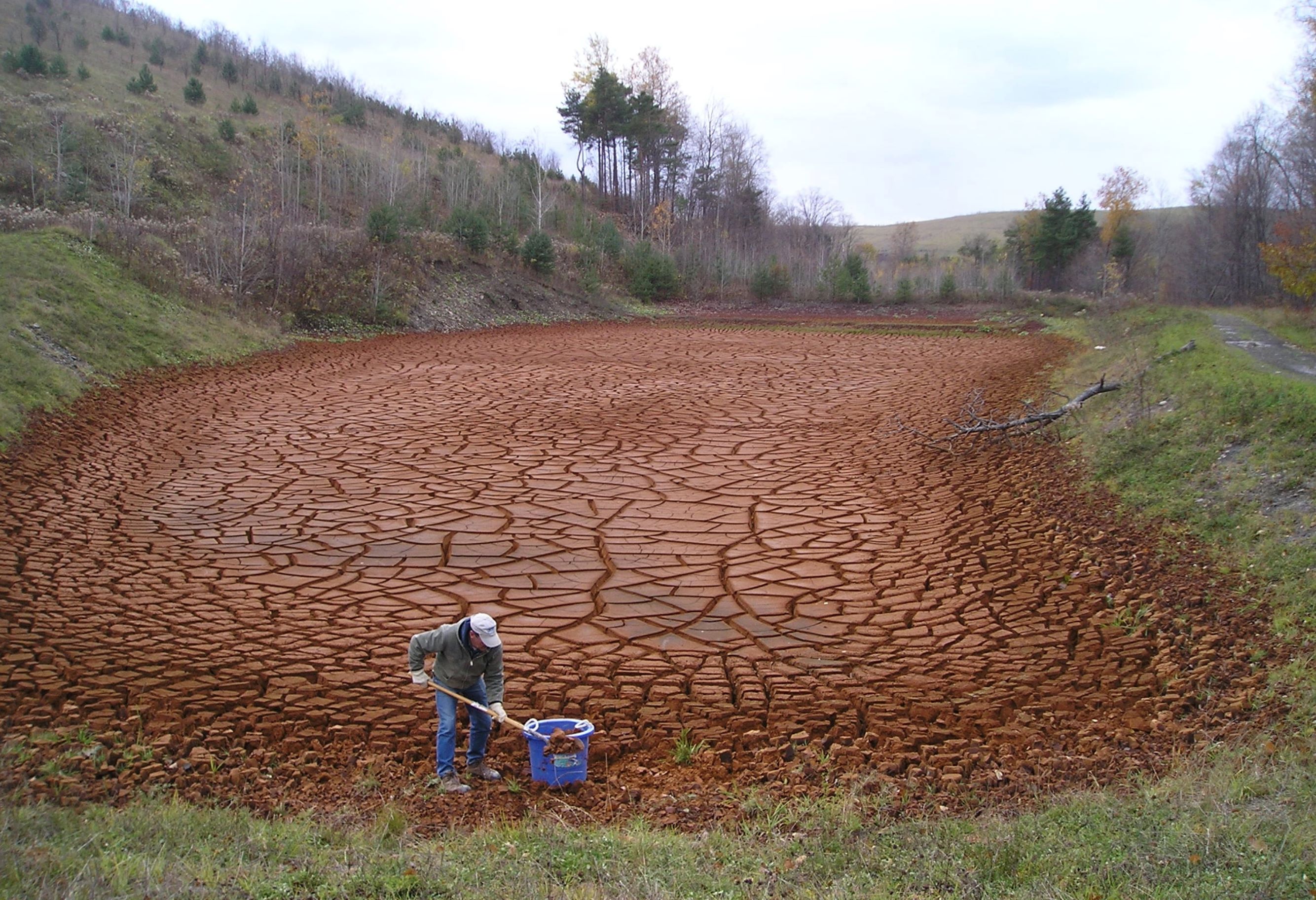 A photo shows a person shoveling acid mine drainage into a bucket while standing on an area of bare, brown, cracked ground, where the drainage has occurred. There is grass around the edges of the drainage area with hills and trees farther away.
