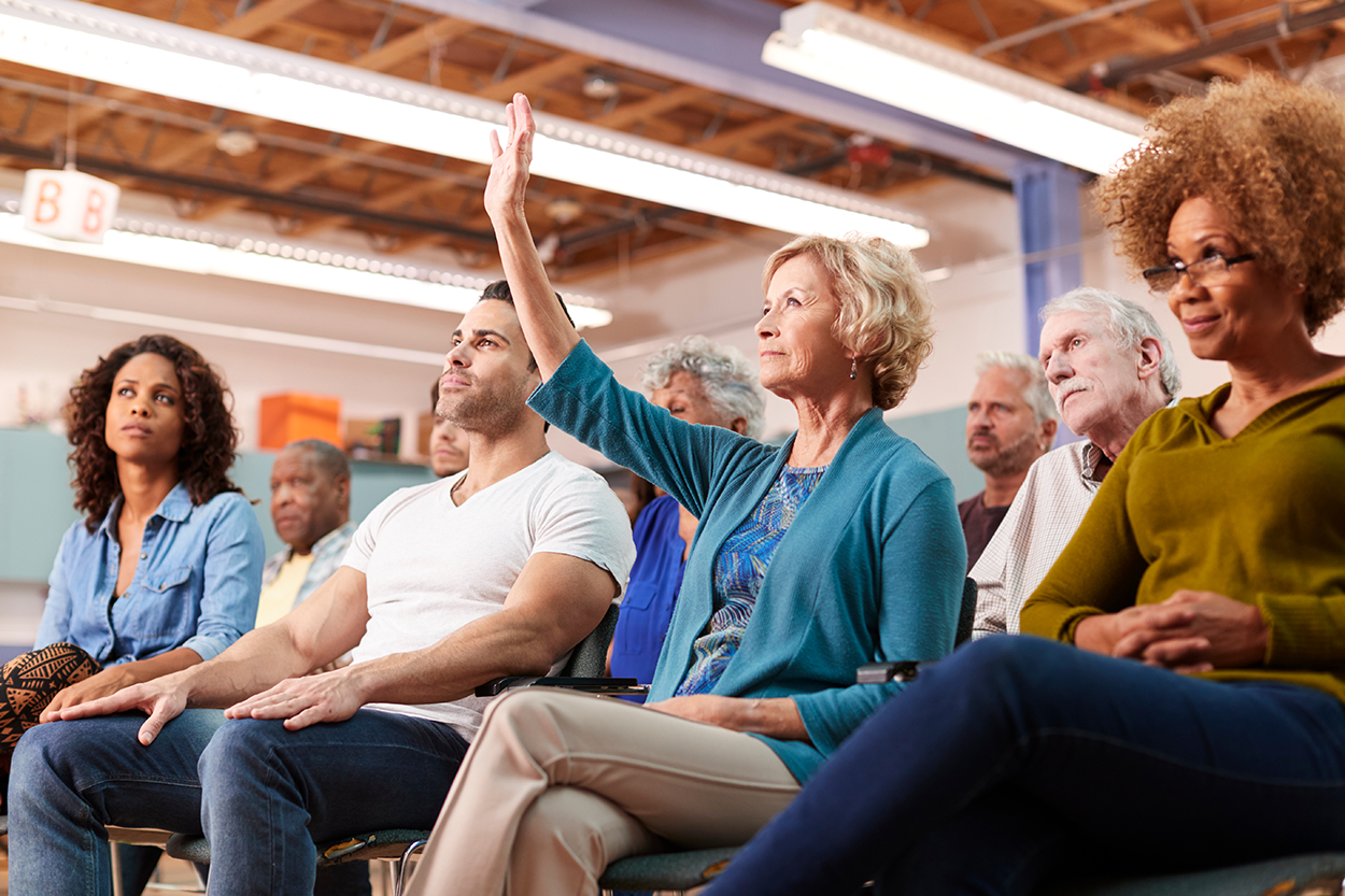 A group of people seated in a meeting room, with one woman raising her hand to speak.