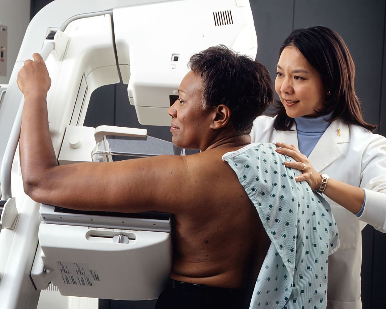 A woman rests one breast on a machine with her arm bent at a right angle to the left. A clinician is nearby helping to position her.